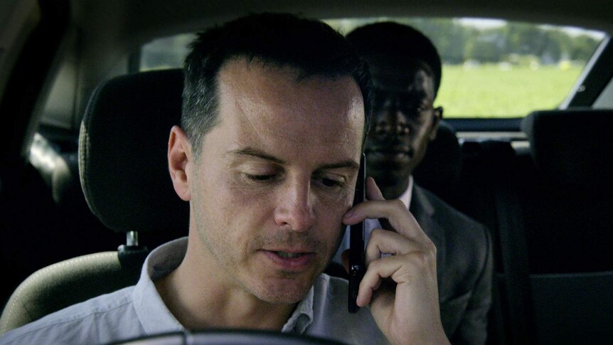 Two men sit in a car, the first in the drivers seat with a phone held to his ear, the second in the back seat looking anxious.