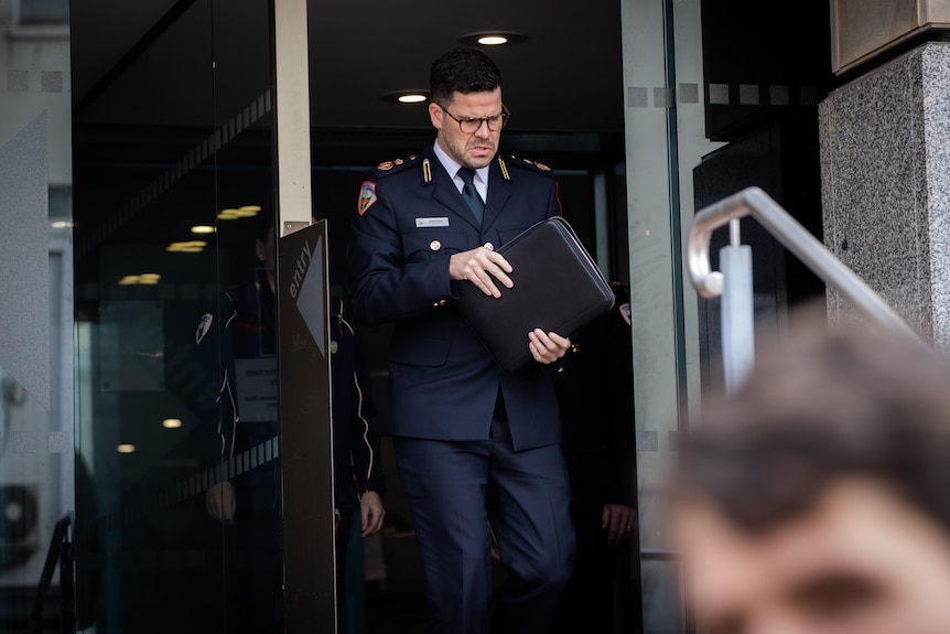 A dark-haired man with glasses and blue uniform walks out of a doorway carrying a folder.