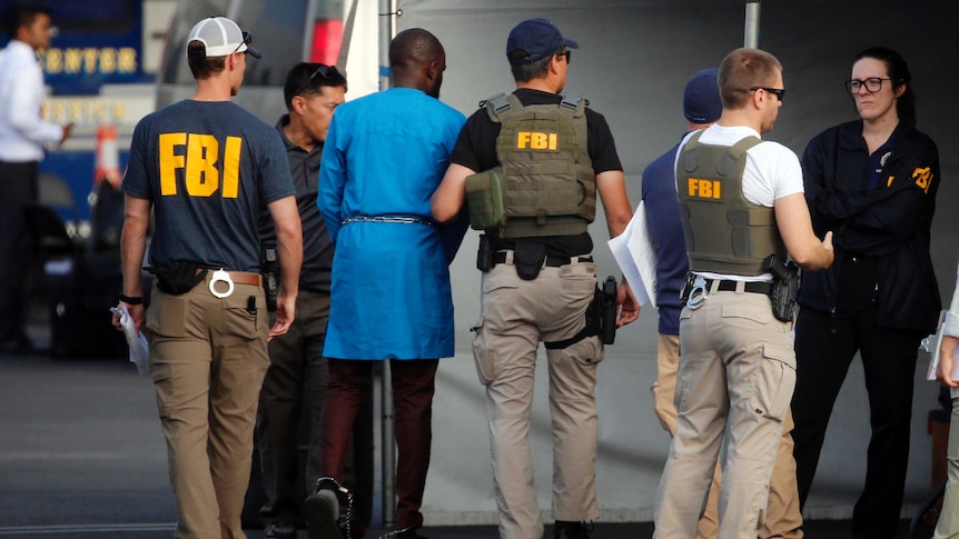 With their backs to the camera, armed FBI agents escort one of the alleged Nigerian nationals fraudsters into a van.