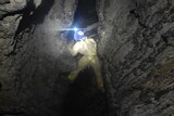 A man with a head torch climbs a rocky wall in a cave.