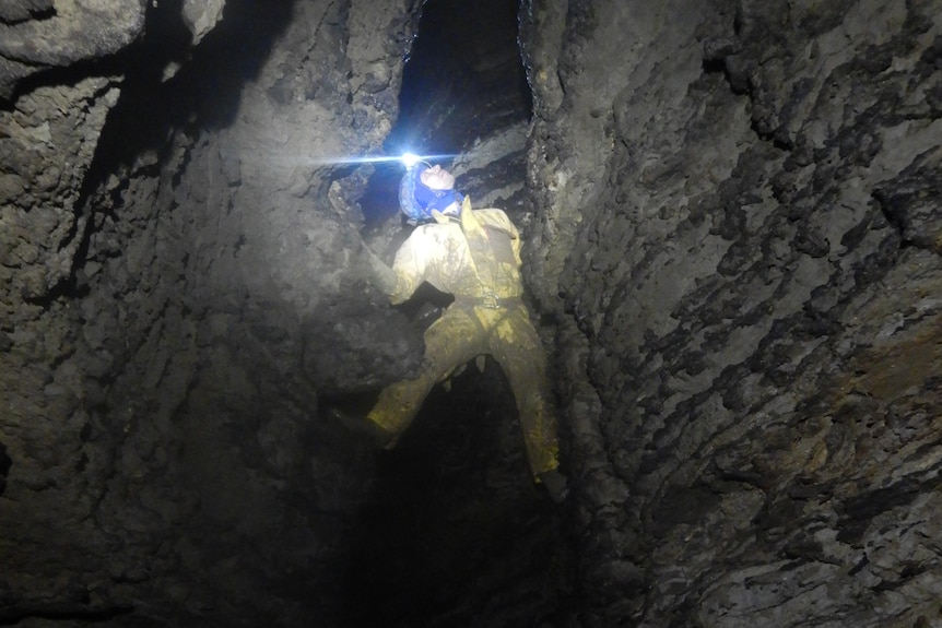 Човек с фенерче се изкачва по скалиста стена в пещера.