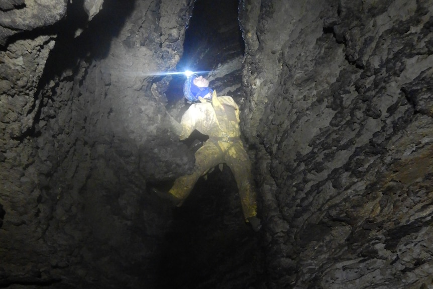 A man with a head torch climbs a rocky wall in a cave.