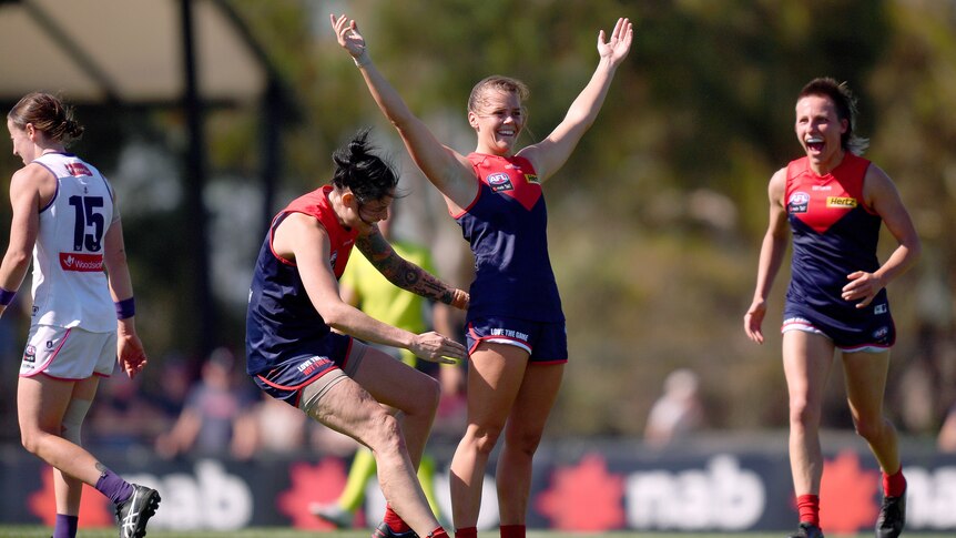 An AFLW player pumps her fist in celebration after a goal as her teammates respond around her.