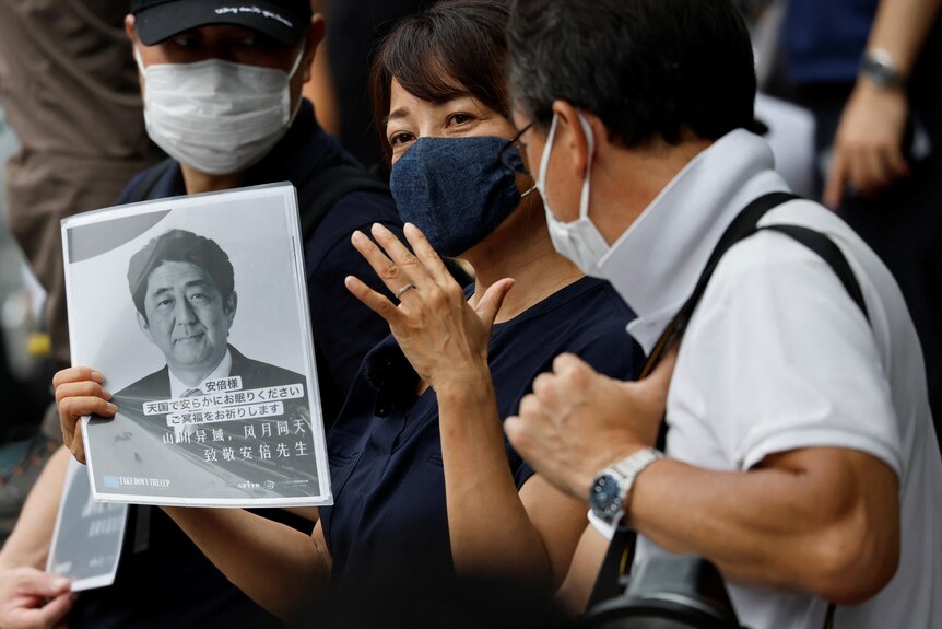 A masked woman in blue holds a photo of Shinzo Abe with Japanese writing below flanked by two masked men