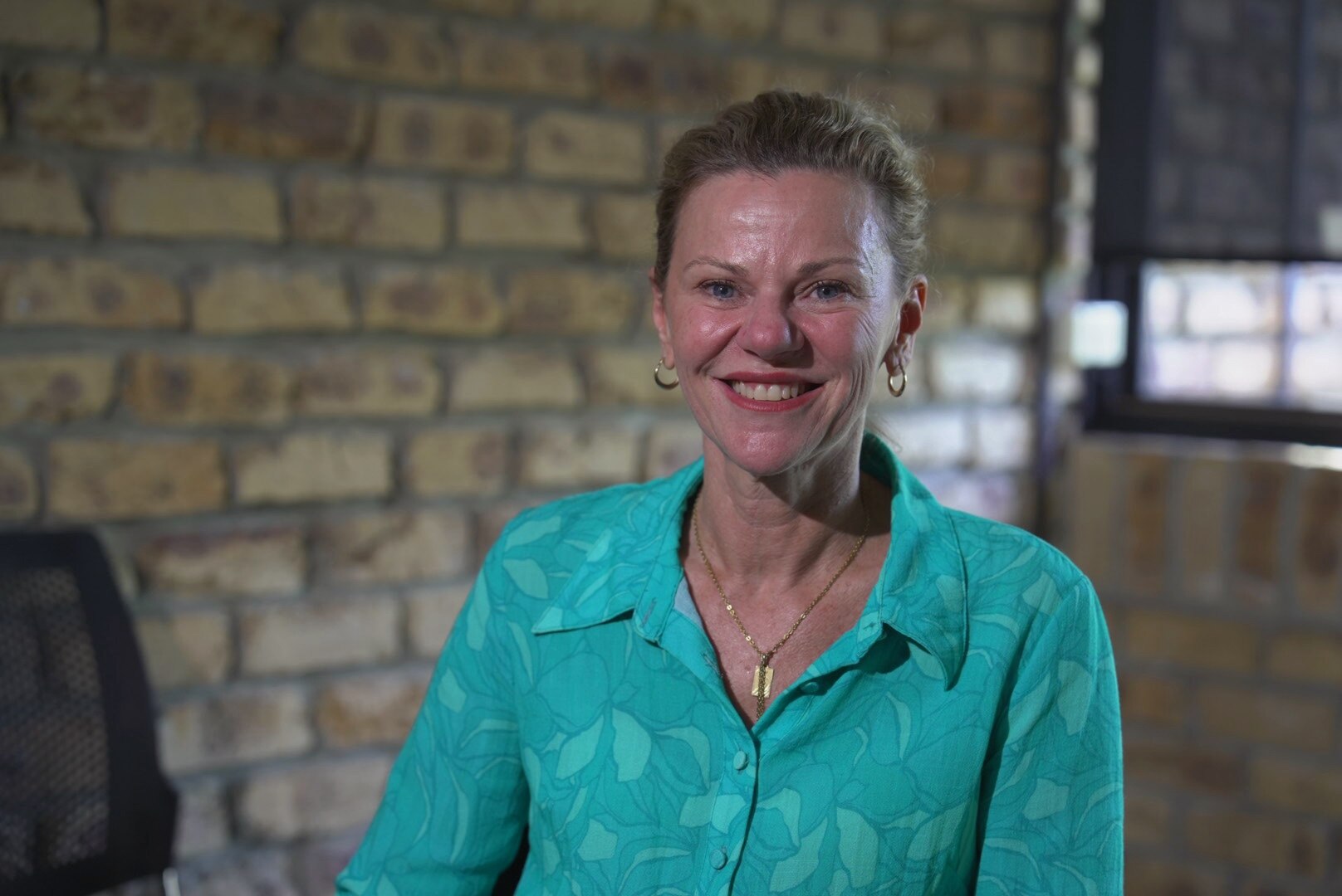 A woman smiles wearing a long sleeve teal blouse in front of a brick wall.