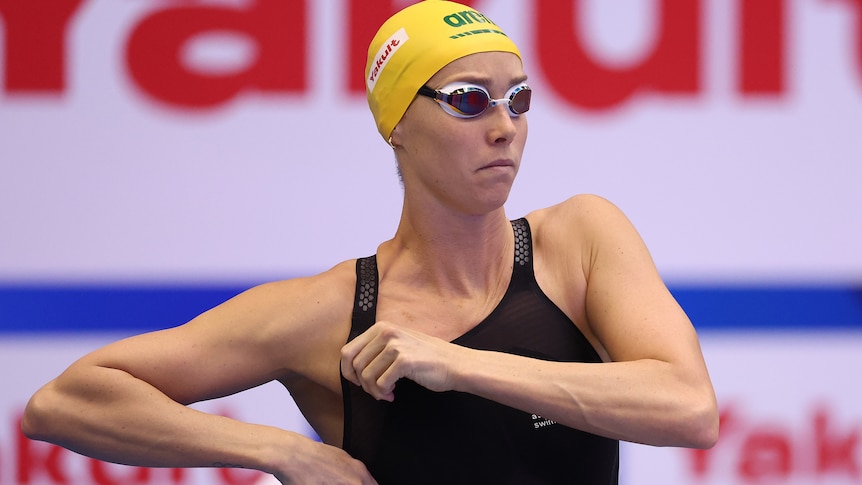 Australian swimmer Emma McKeon adjusts her swimsuit before a race. She is wearing a cap and goggles.