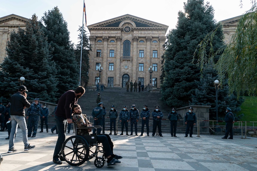 A soldier in a wheelchair sits outside an official building with a line of police in front of him.