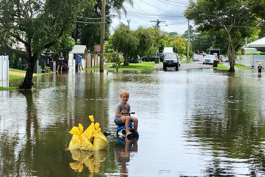 A boy sits on a trike in floodwater with three sandbags next to him.