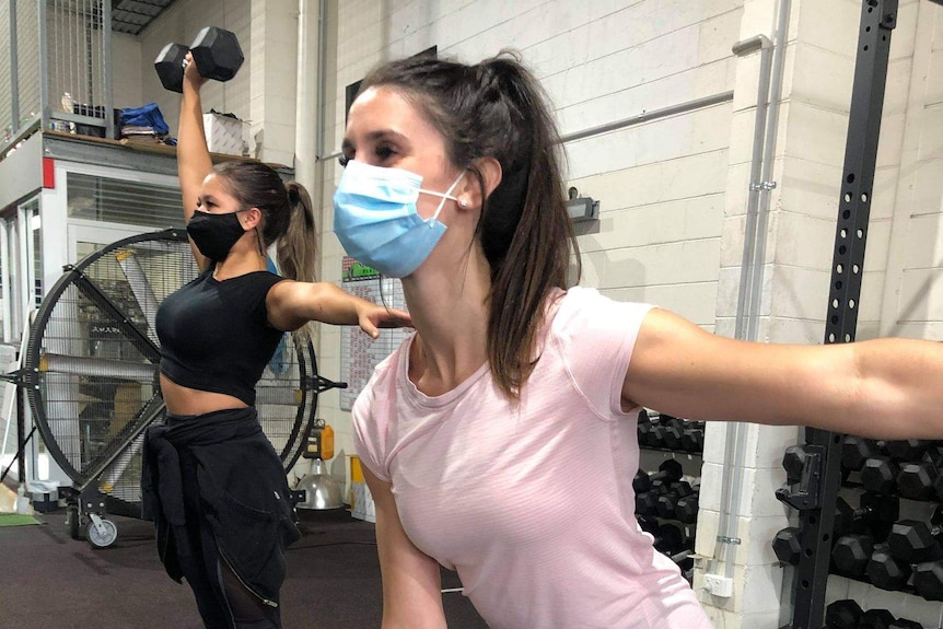 Two women work out with dumb bells and masks on.