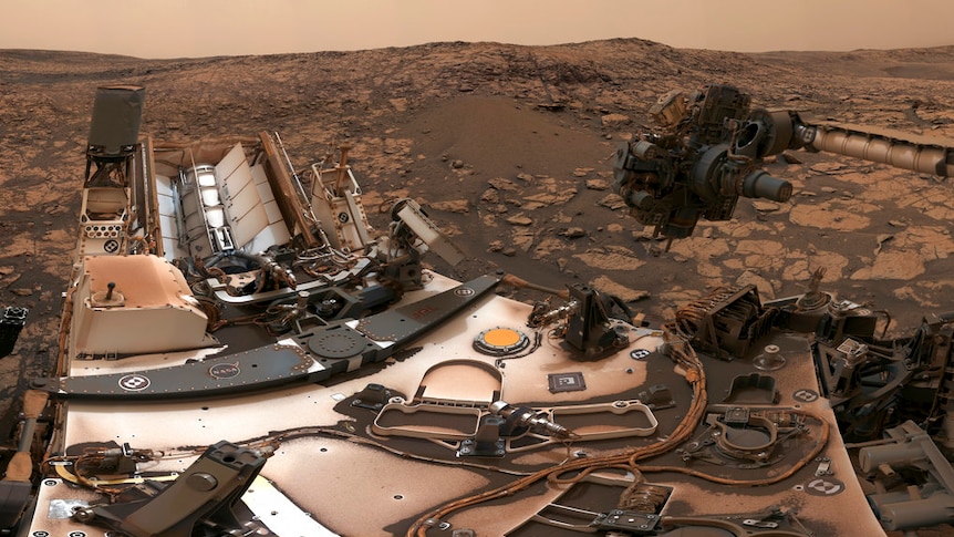 A dusty rover with a camera on a mast above it among red rocks, under a darkened reddish-brown sky