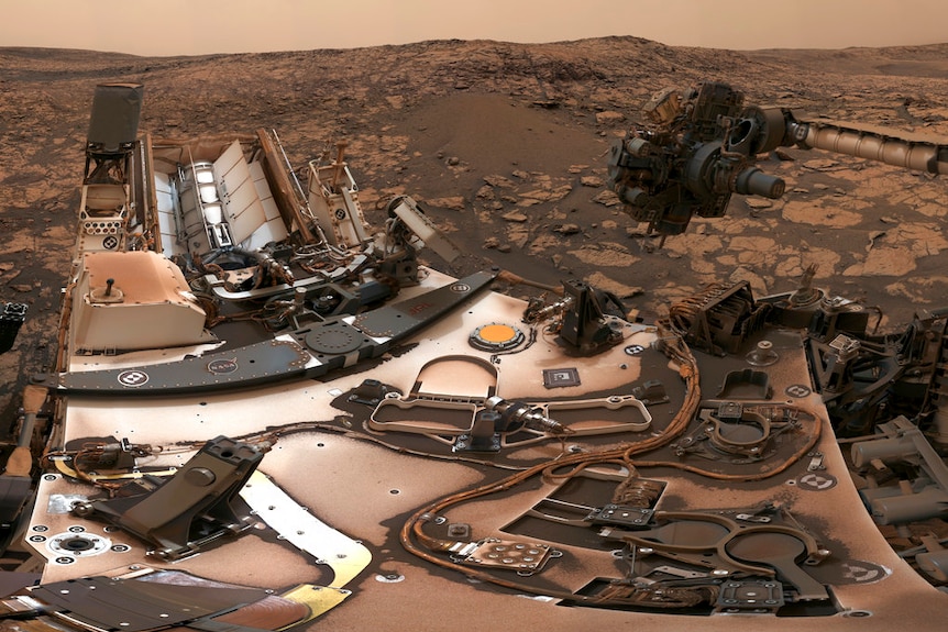 A dusty rover with a camera on a mast above it among red rocks, under a darkened reddish-brown sky