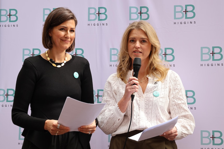 Two white women stand while one holds a microphone, giving a speech