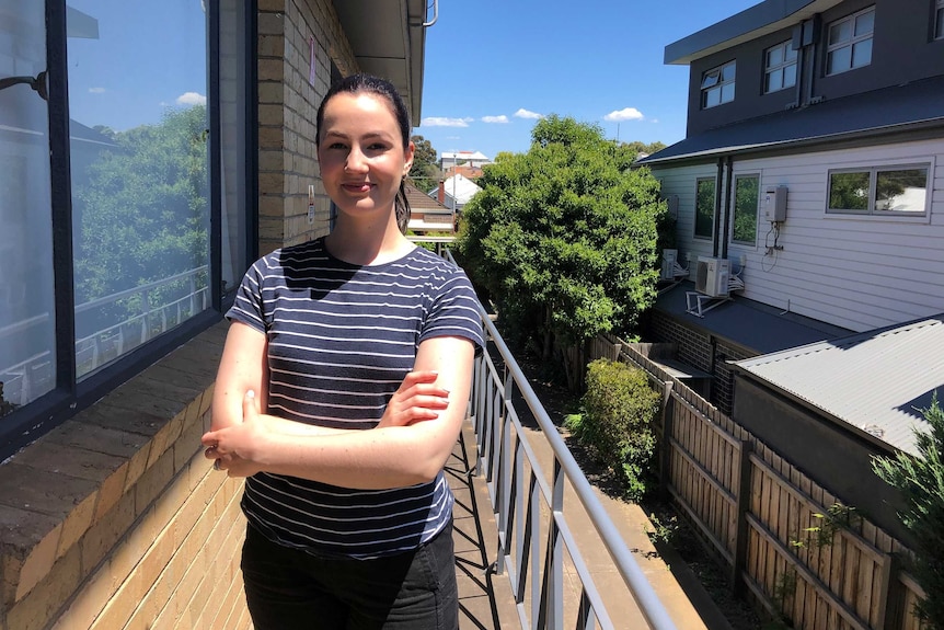 A young woman wearing a black and white striped t-shirt stands outside her apartment, with her arms folded smiling to the camera