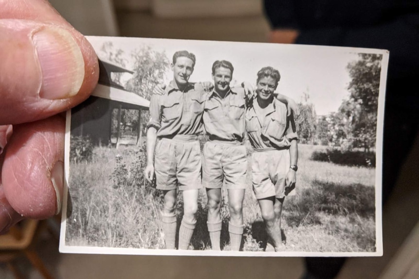 Frank Sims holds a small black and white photo of himself and two other soldiers with their arms around each other's shoulders.