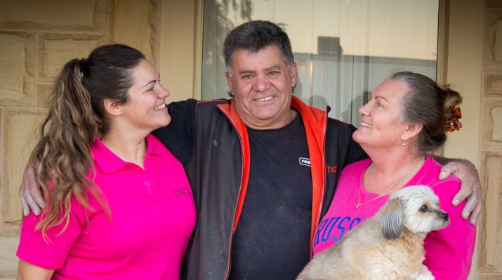 Mick Albanese stands in front of a house window with his daughter and wife, who is holding a small pet dog.