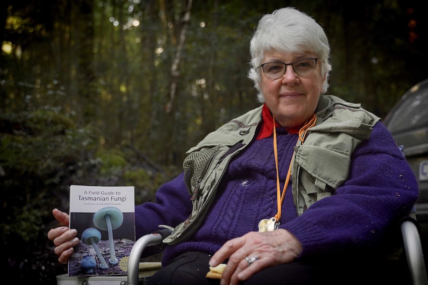 Pat Harrison sitting outside in a foldup chair, wearing a purple jumper and holding a copy of Field Guide to Tasmanian Fungi