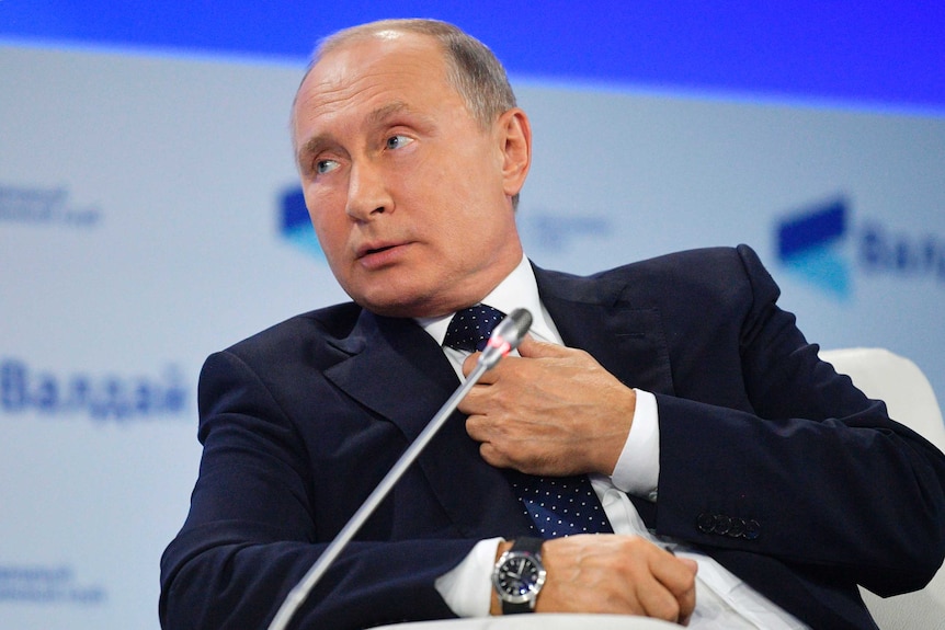 Russian President Vladimir Putin adjusts his tie and looks to the left whilst seated speaking at a meeting.