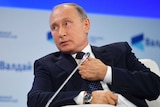 Russian President Vladimir Putin adjusts his tie and looks to the left whilst seated speaking at a meeting.