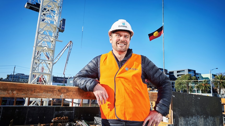 A man in a high-vis vest and hard hat with Aboriginal flag behind him.