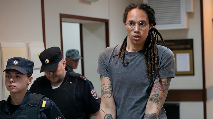 A tall, tattooed woman with dreadlocks is led, handcuffed, down a hallway by two guards wearing blue caps and uniforms.