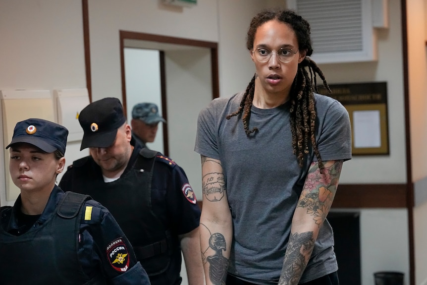 A tall, tattooed woman with dreadlocks is led, handcuffed, down a hallway by two guards wearing blue caps and uniforms.