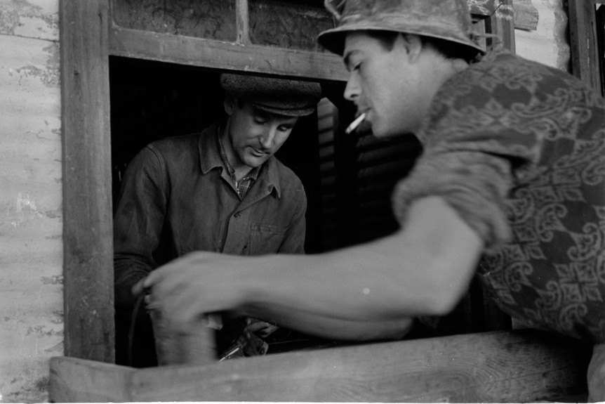 A black and white photo of a man wearing a hat standing outside, speaking to another man through a window