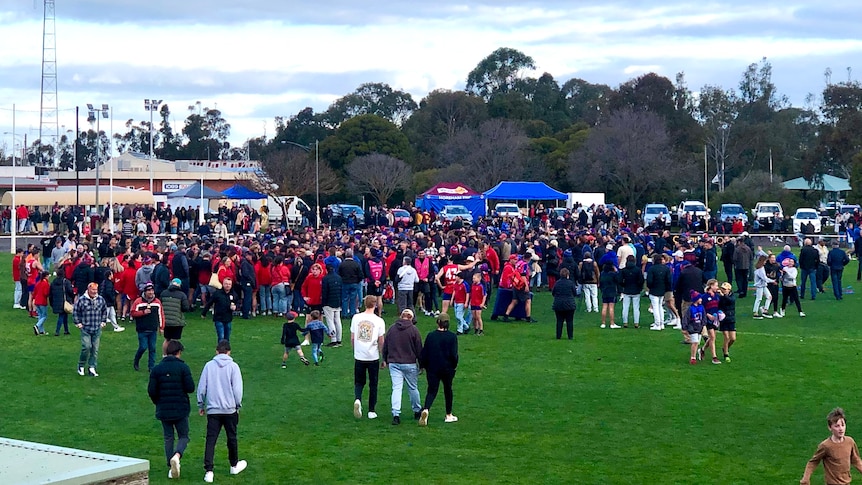 A crowd of footy fans many dressed in red congregate at a footy oval with stands in the back
