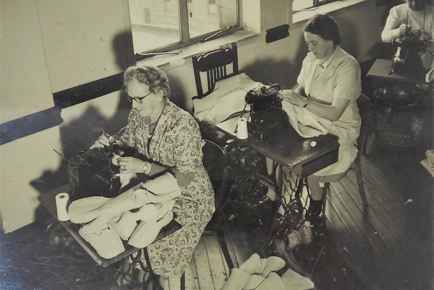 An old black and white photograph of three older CWA women sitting at tables sewing sheepskin vests.