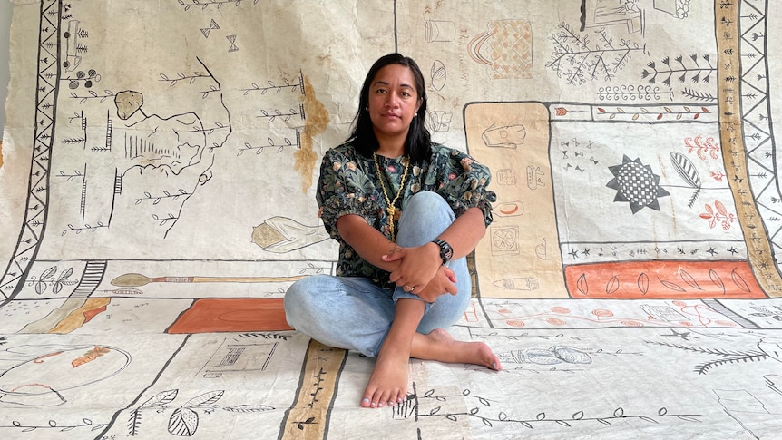 A woman with black hair sits with her legs crossed on a traditional Niuean bark cloth mat with patterns.