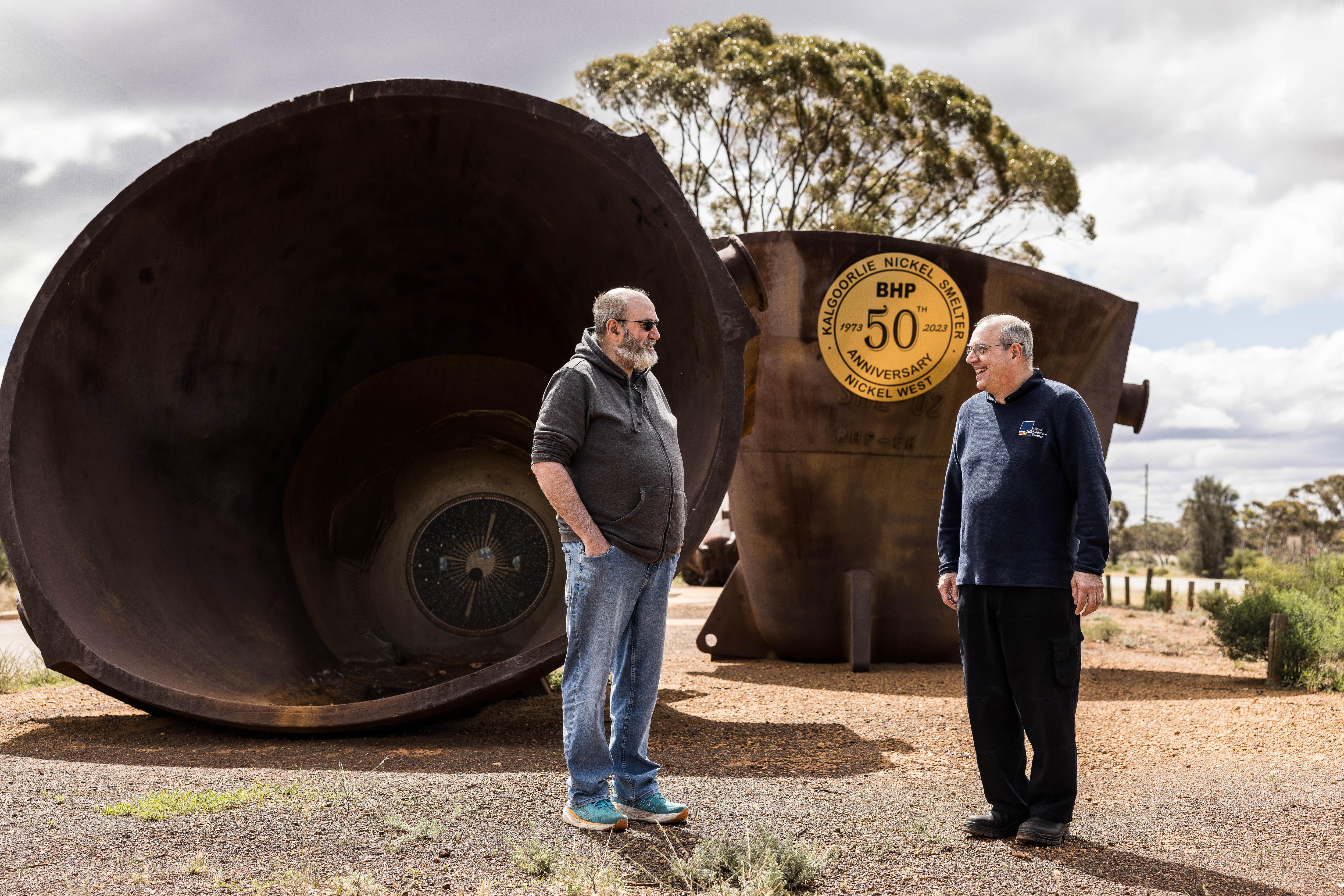 Two men standing in front of large industrial-scale pots used in a metal furnace.  