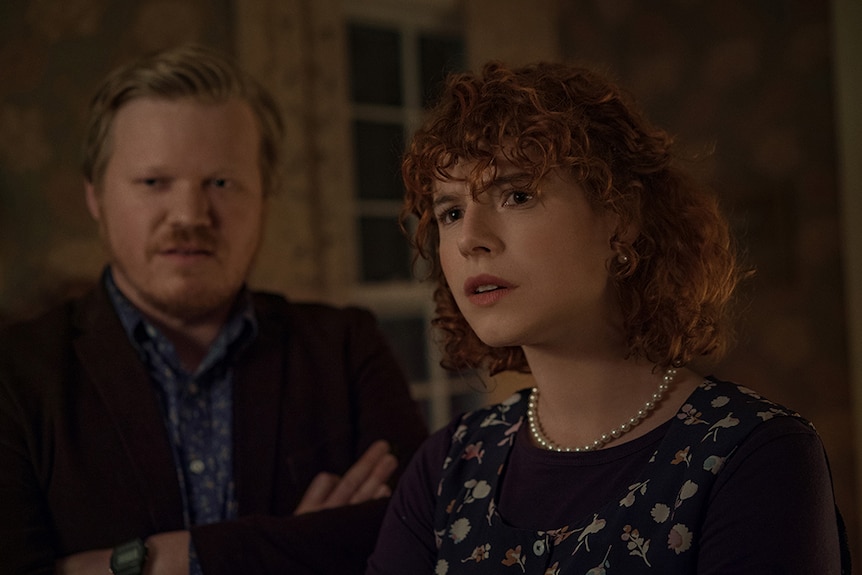 A blonde bearded man with arms folded and woman with curly red hair stand in interior room look towards something out of frame.