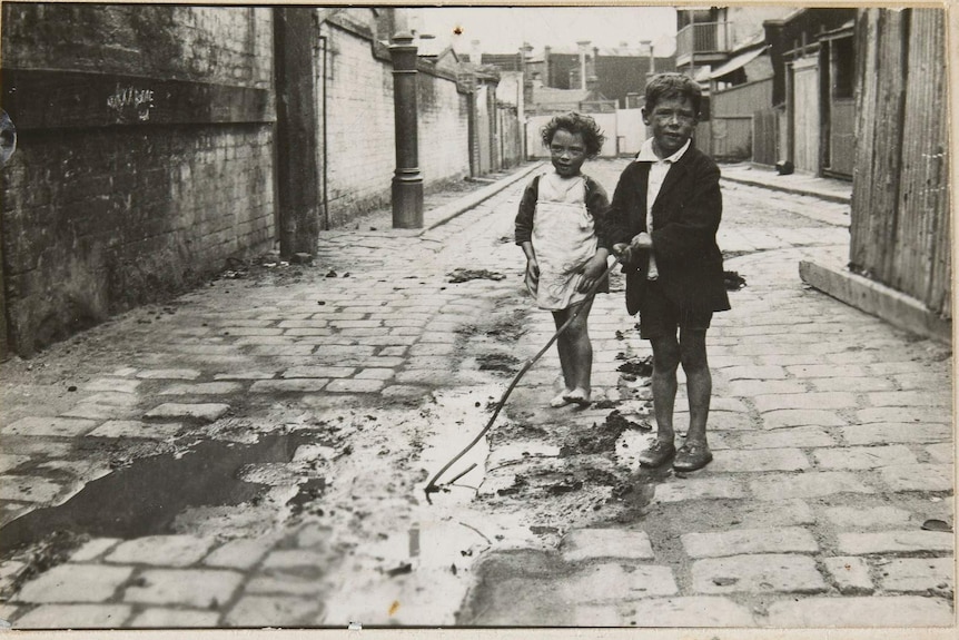 Two dirty children playing in a pool of water in a street paved with bluestone. Houses visible in the background.