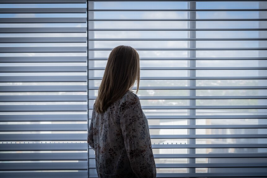 A woman stands with her back turned looking outside white vertical blinds