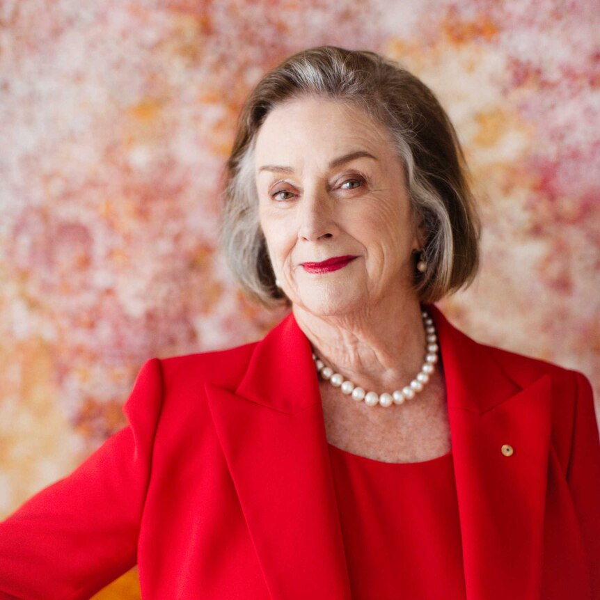 Wendy McCarthy in a red suit against an abstract rose and orange background