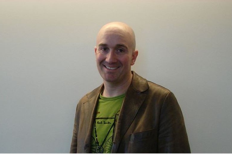 Simon Kennedy, bald, in tan jacket with green shirt, smiles, standing against a white wall.