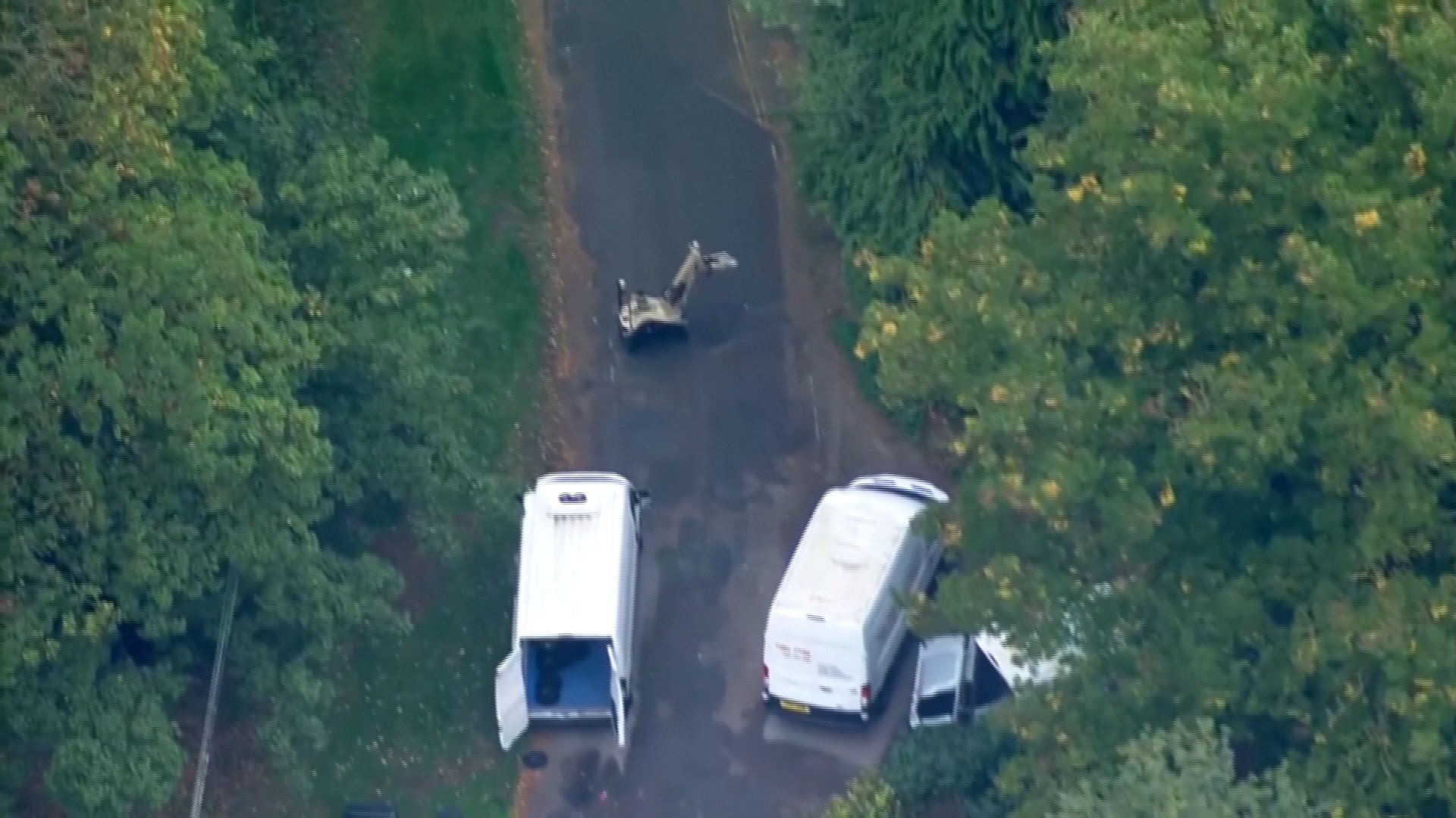 Two white vans, with one of its rear doors open, seen on a roadway in between green trees and a tan bomb disposal robot.