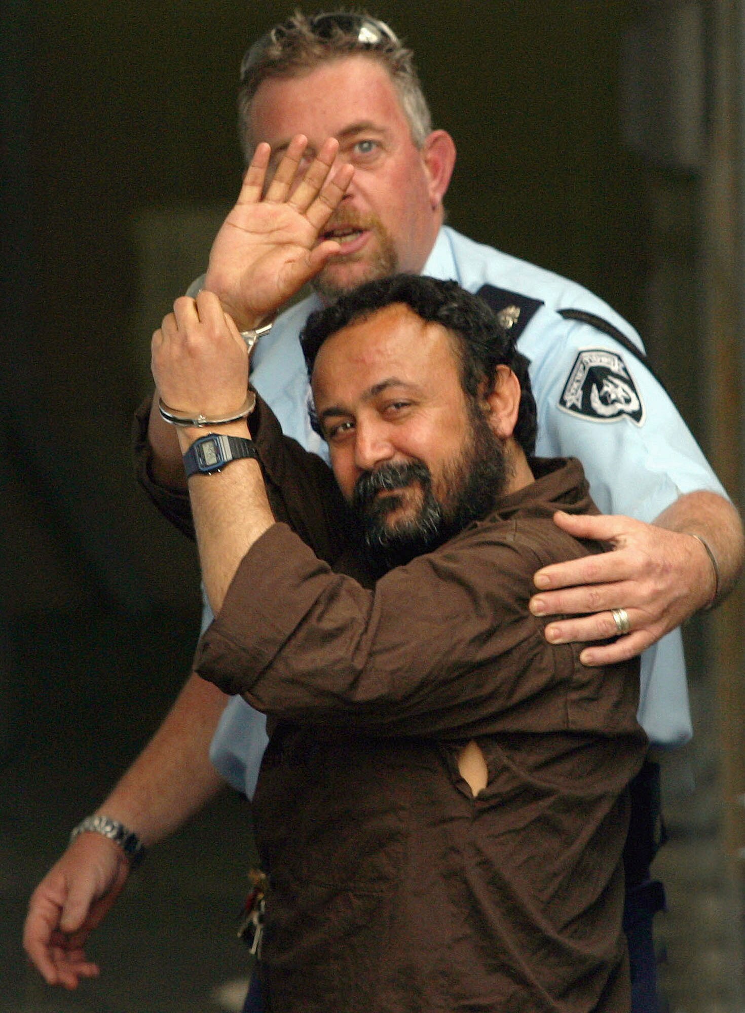 A security guard leads Marwan Barghouti, who is smiling and waving, into a court.