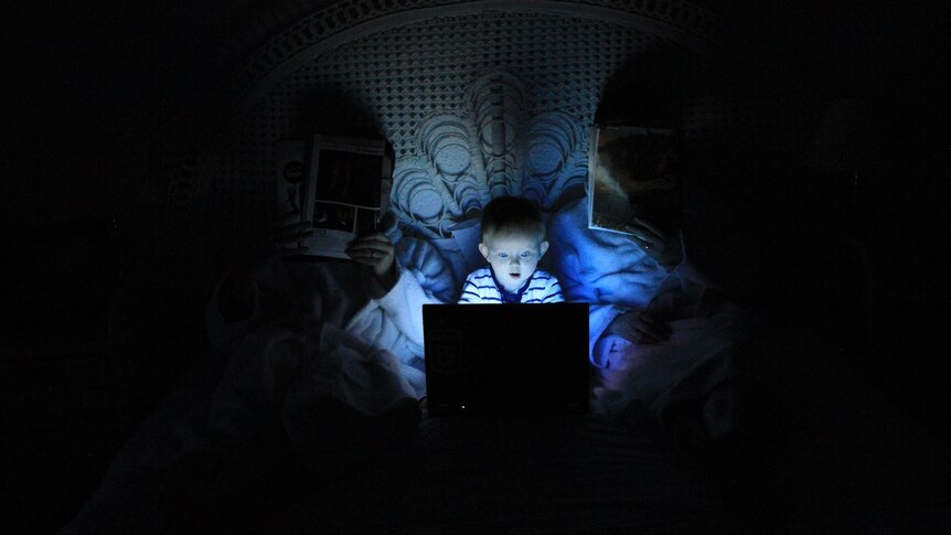 A small child in bed with parents. The child is lit by the glow of a laptop screen, while the parents have newspapers.