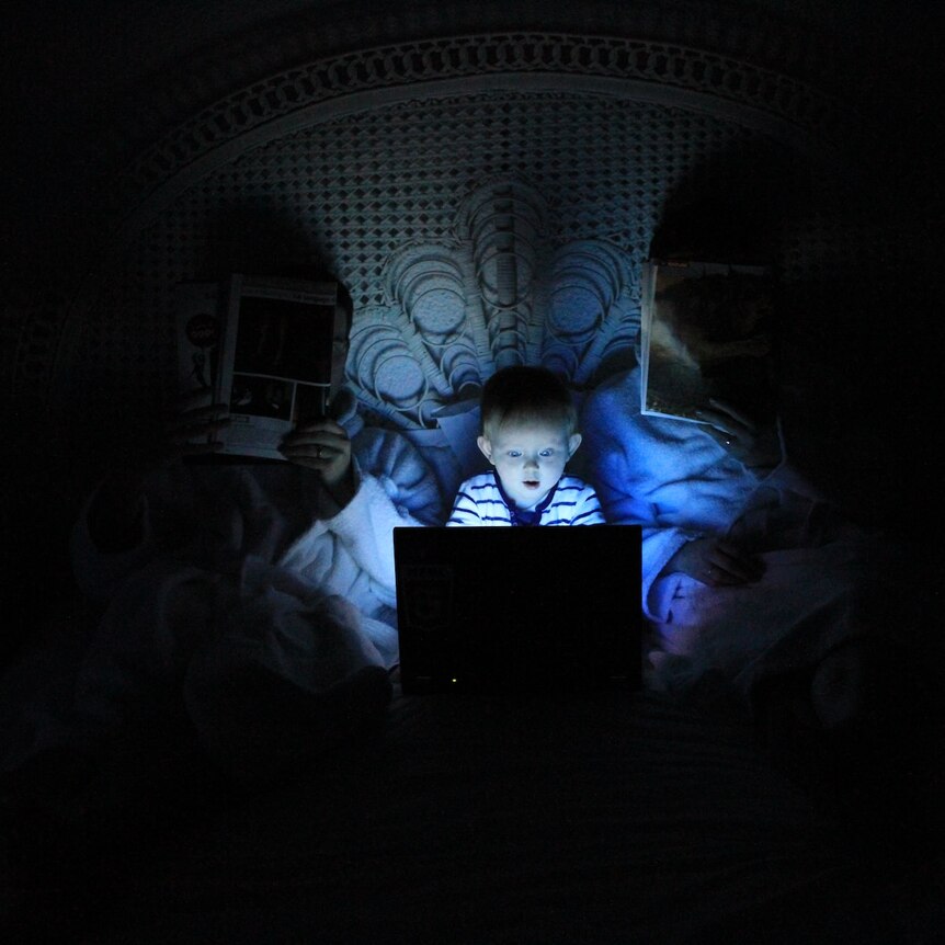 A small child in bed with parents. The child is lit by the glow of a laptop screen, while the parents have newspapers.