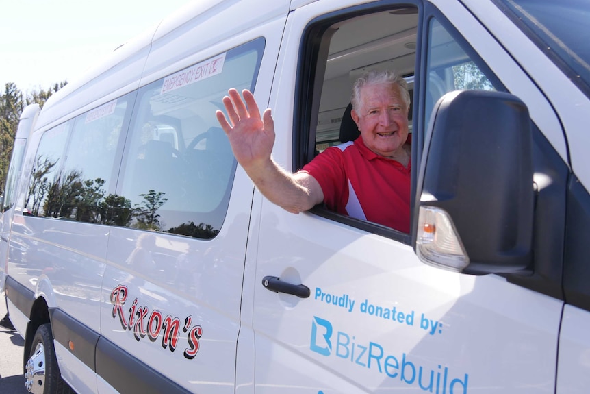 Mal Rixon sits in the drivers seat of the mini bus wearing a red shirt. He is waving with a smile of his face.