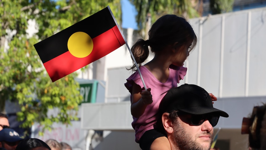 A young woman is on the shoulders of a man in a march. She's holding an Aboriginal flag