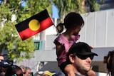 A young woman is on the shoulders of a man in a march. She's holding an Aboriginal flag
