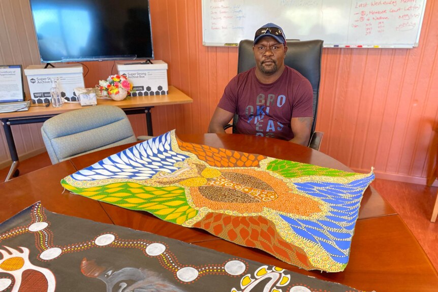 A man sits at a table. In front of him is intricate and colourful Indigenous art 