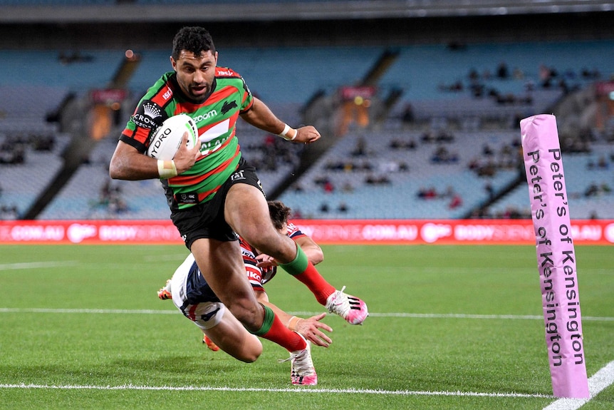 A South Sydney NRL players holds the ball with his right arm as he crosses the line for a try against the Roosters.