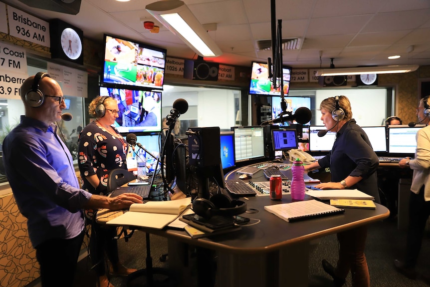 Two broadcasters standing at desks wearing headphones and talking into microphones.