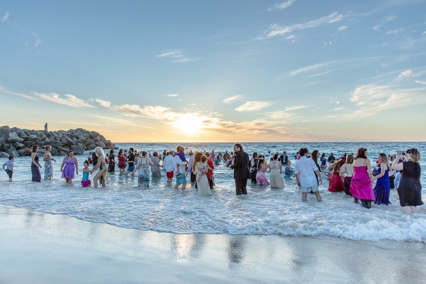A large crowd of people in formal wear swim in the ocean at sunset.