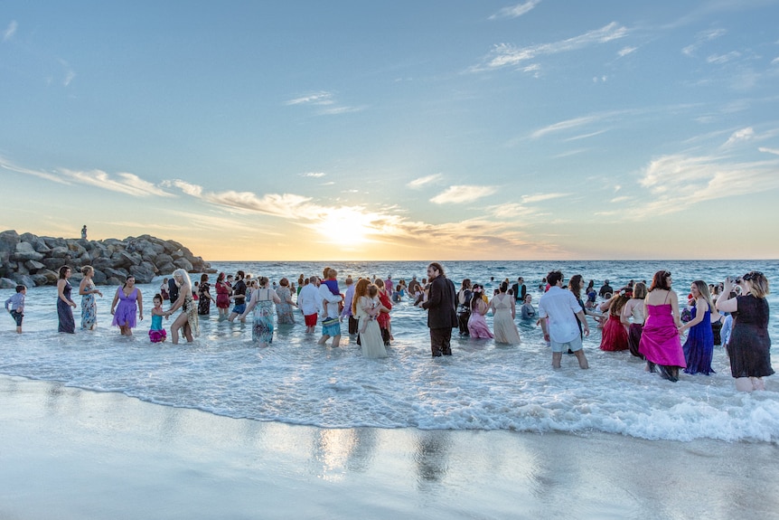 A large crowd of people in formal wear swim in the ocean at sunset.