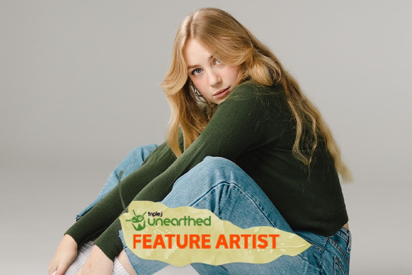 Kat Edwards, a young woman with long strawberry blonde hair, a green shirt and jeans, sits and looks toward the camera.