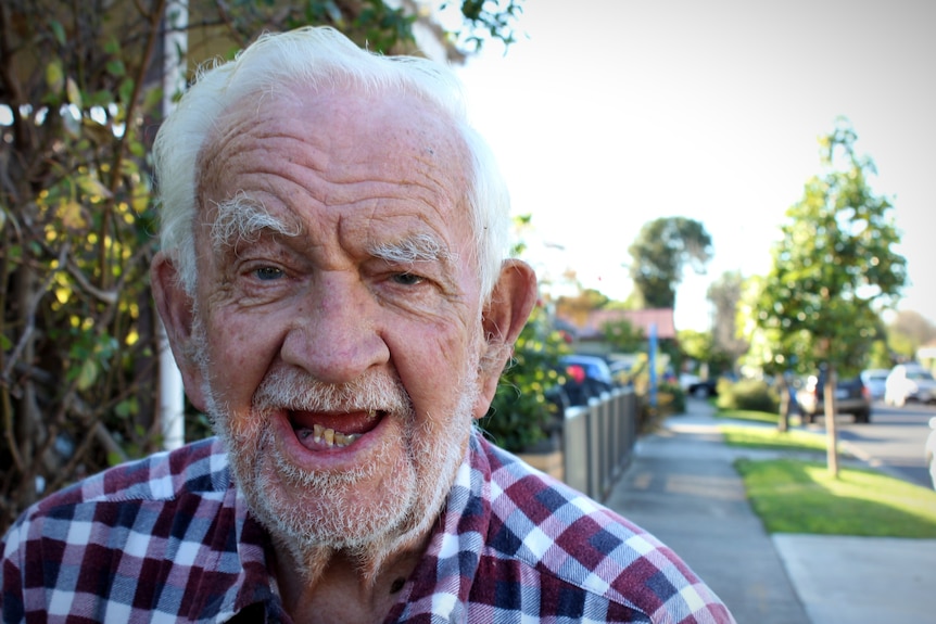 A close up of an elderly man with a checked shirt standing on a suburban street.