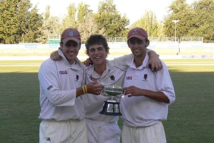 Three men holding a trophy while dressed in cricket uniform.